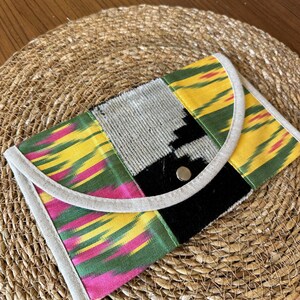 Peut inclure: Une pochette colorée avec un design patchwork. Le tissu présente des motifs jaunes, verts, roses et noirs. La pochette a un rabat incurvé avec une bordure de couleur neutre et une fermeture à bouton-pression en métal. Sur un tapis tissé.