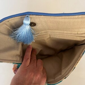 May include: A beige fabric clutch bag with a blue trim and a blue tassel. The bag is open and the inside is visible.