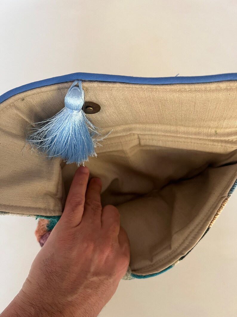 May include: A beige fabric clutch bag with a blue trim and a blue tassel. The bag is open and the inside is visible.