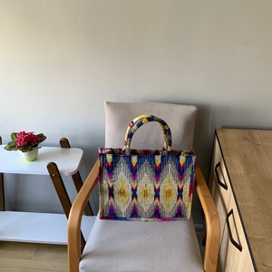 May include: A colorful tote bag with a geometric pattern in shades of blue, yellow, pink, and gray. The bag has a brown leather handle and is sitting on a light brown chair.