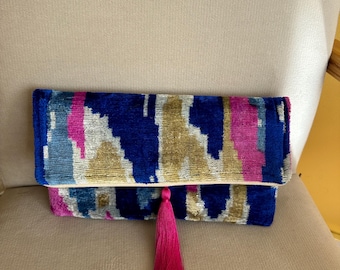 May include: A blue and gold clutch purse with a pink tassel. The clutch has a geometric pattern and is made of a soft, textured fabric.