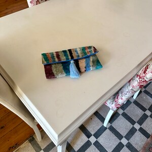 May include: A colorful striped clutch with a blue tassel on a white table. The clutch is made of a soft, textured fabric and has a blue trim.