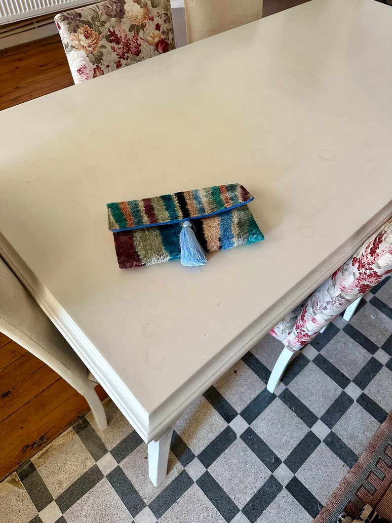 May include: A colorful striped clutch with a blue tassel on a white table. The clutch is made of a soft, textured fabric and has a blue trim.