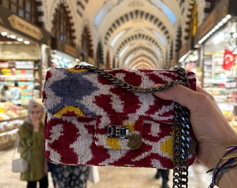 May include: A red, white, and blue patterned velvet handbag with a silver chain strap. The bag has a silver clasp and is held in a person's hand.