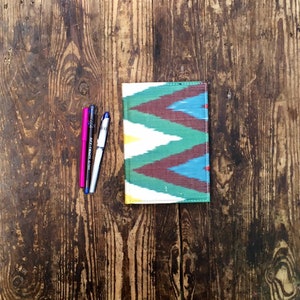 May include: A colourful notebook with a geometric pattern in shades of blue, green, red, and white. The notebook is lying on a wooden surface with three pens next to it.
