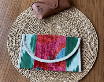 May include: A small, rectangular, multi-coloured fabric pouch with a flap closure. The pouch has a green, white, and blue striped pattern on the outside and a red, pink, and blue patterned fabric on the inside. The pouch is sitting on a woven straw mat.