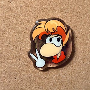May include: A clear acrylic pin featuring a cartoon character with orange hair, a large nose, and a red shirt. The character is making a peace sign with their hand. The pin is set against a brown background.