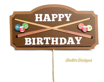 May include: A brown and tan birthday cake topper with the words "HAPPY BIRTHDAY" in white. The topper features two crossed pool cues, and three pool balls with the number 11. The topper is on a wooden stick.