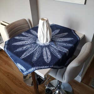 May include: A white ceramic vase with a textured surface sits on a blue and white table runner. The table runner has a circular design with white accents. The table is white and has a square shape. Two gray chairs with chrome legs are visible in the background.