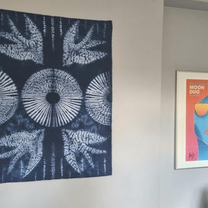 May include: A dark blue and white textile wall hanging with a symmetrical pattern of circles and leaf-like shapes. A framed poster with the text "MOON DUO" and a colourful abstract face is also visible.