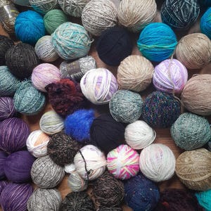 May include: A selection of yarn balls in a variety of colours including blue, green, purple, white, black, and brown. The balls of yarn are tightly packed together, displaying diverse textures and hues, suitable for knitting and crochet.