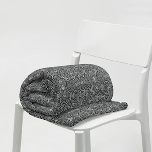 Può includere: Una coperta arrotolata con un motivo geometrico grigio e bianco, appoggiata su una sedia bianca. Il motivo della coperta presenta una complessa disposizione di linee e forme. La coperta è arrotolata, mostrando il suo motivo e la sua consistenza.