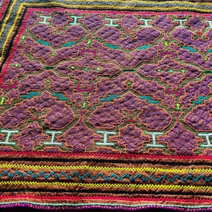 Può includere: Un arazzo tessuto colorato con un motivo geometrico in tonalità di viola, rosso, verde e giallo. L'arazzo ha un bordo marrone con cuciture intricate.