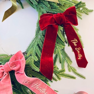 May include: A festive Christmas wreath featuring green foliage and three velvet bows in red, pink, and olive green. Each bow is personalised with a family name in gold lettering: "The Smiths", "The Heaths", and "The Holmes".