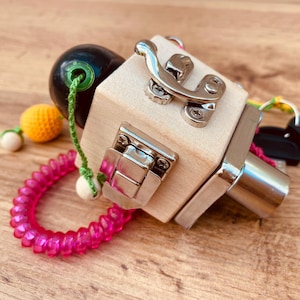 Puede incluir: Un cubo de madera con varios cierres, un resorte, una bola y una llave. El cubo está hecho de madera natural y tiene un color marrón claro. Los cierres son de metal y tienen un acabado plateado. El resorte es rosa y está unido al cubo con una cuerda verde. La bola es negra y tiene una cuerda verde atada a ella. La llave es plateada y tiene un mango negro.