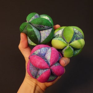 Peut inclure: Trois boules en tissu, une verte, une rose et une vert clair, sont tenues dans une main. Les boules sont faites de carrés de tissu cousus ensemble pour former une sphère.