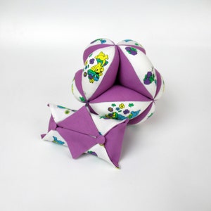 Peut inclure: Une balle en tissu violet et blanc avec un motif de dessin animé coloré. La balle est composée de plusieurs panneaux de tissu cousus ensemble. Une petite pochette en tissu violet est également incluse.