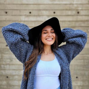 May include: A woman wearing a black wide-brimmed hat and a blue knit cardigan. The cardigan has a textured appearance and is paired with a white tank top. The background is a textured concrete wall.