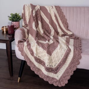 May include: A handmade, crocheted blanket in shades of mauve and cream, draped over a pink sofa. The blanket features a scalloped edge and a geometric pattern. A small side table with a plant and a drink is visible.