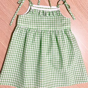 Puede incluir: Un vestido de verano a cuadros verdes y blancos con tirantes anudados. El vestido presenta un escote con volantes y una falda fruncida. La tela es de algodón ligero. El vestido está diseñado para un niño.