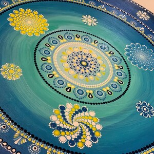 May include: A blue and green mandala with yellow and white accents. The mandala is painted on a round canvas and features a circular pattern of dots and swirls.