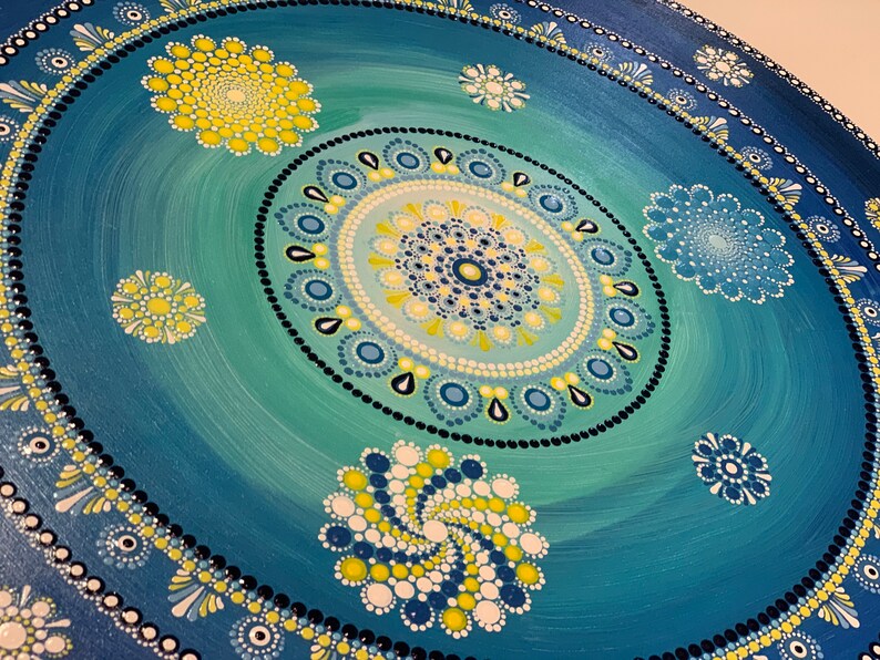 May include: A blue and green mandala with yellow and white accents. The mandala is painted on a round canvas and features a circular pattern of dots and swirls.
