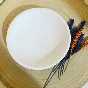 Peut inclure: Un plat rond et blanc, posé sur une base en bois, est orné de fleurs séchées bleues et oranges. Le plat a une surface lisse et semble être en céramique. Le fond en bois présente des motifs naturels.