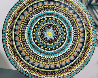 May include: A circular mandala design painted on a wooden surface. The mandala features concentric circles of dots in various colours, including blue, green, brown, and white.