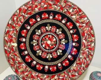 May include: A black circular plate with a red, white and brown mandala design. The mandala is made up of dots and swirls. The plate is on a wooden easel.