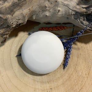 May include: A white, round, ceramic soap dish sits on a light brown, wooden surface. The soap dish is surrounded by dried blue and orange floral elements and a piece of driftwood.