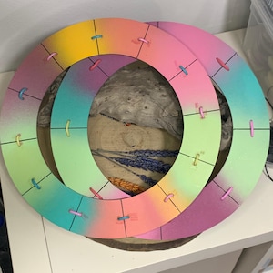 Pode incluir: Duas molduras circulares de madeira sobrepostas com um gradiente de cores, incluindo amarelo, rosa, verde e azul. Cada moldura tem pequenas costuras coloridas espaçadas uniformemente. As molduras são provavelmente para artesanato ou decoração de casa.