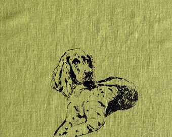 May include: A light green linen fabric with a black illustration of a dog. The dog is lying down with its head up and ears flopped over. The illustration is detailed and the fabric has a textured appearance.