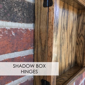 May include: Close-up of two black metal hinges attached to a wooden shadow box. The box is made of stained wood with a visible wood grain. The text "SHADOW BOX HINGES" is printed on a white label.