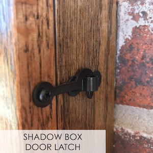 May include: Close-up of a black metal door latch attached to a wooden door. The latch is in the shape of a hook and has a small screw on the side. The text "SHADOW BOX DOOR LATCH" is visible below the latch.