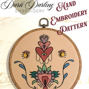 May include: Embroidered hoop art with a pink, red, and green floral design. The design features a stylized heart with an eye in the center, surrounded by smaller hearts. The hoop is made of wood and has a brass ring.