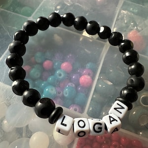 May include: Black beaded bracelet with white letter beads spelling out the name "LOGAN".