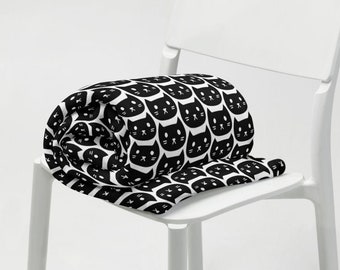 May include: A rolled-up black and white throw blanket with a repeating pattern of cat faces. The blanket is draped over the seat of a white chair. The cat faces have black outlines, eyes, and noses, with white backgrounds.
