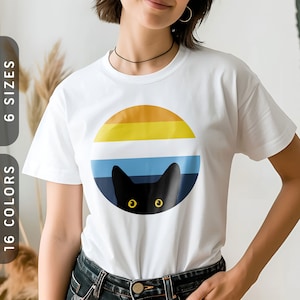 May include: White t-shirt featuring a black cat peeking out from behind a colourful sunset graphic. The graphic is a circle with stripes of orange, yellow, blue, and white.