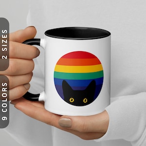 May include: White ceramic mug with a black rim and handle. The mug features a rainbow circle with a black cat silhouette peeking out from the bottom.
