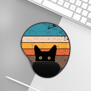 Può includere: Un tappetino per mouse rotondo nero con un design retrò che presenta un gatto nero con occhi gialli che spuntano da uno sfondo a strisce multicolore.