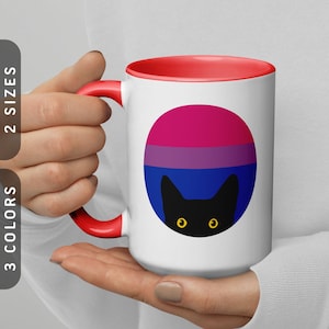 Puede incluir: Taza de cerámica blanca con borde rojo. La taza tiene una silueta de gato negro con ojos amarillos que asoman detrás de una bandera del orgullo bisexual.