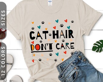 Pode incluir: Uma camiseta bege com o texto "Cat Hair Don't Care" em letras pretas e coloridas. A camiseta é decorada com pequenos corações e pegadas de patas de várias cores.
