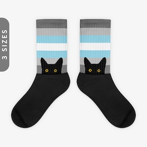 May include: A pair of black socks with a black cat peeking out from the top. The socks feature a grey, white, and light blue striped pattern, resembling the transgender pride flag.