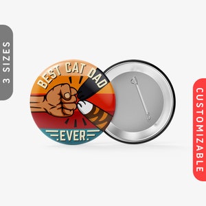 May include: A round button with a retro-style design featuring a fist bump between a human hand and a cat paw. The text "Best Cat Dad Ever" is written in a bold font on the button.