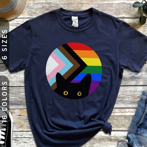May include: A navy blue t-shirt with a black cat peeking out from behind a circle of rainbow and pride flag colors. The colors in the circle are pink, light blue, white, brown, black, and the rainbow colors.