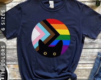 May include: A navy blue t-shirt with a black cat peeking out from behind a circle of rainbow and pride flag colors. The colors in the circle are pink, light blue, white, brown, black, and the rainbow colors.