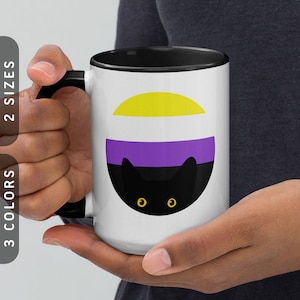 Puede incluir: Taza de cerámica blanca con borde y asa negros. La taza presenta un gato negro con ojos amarillos que asoma detrás de un círculo con los colores de la bandera del orgullo no binario: amarillo, blanco, morado y negro.