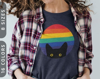 May include: Navy blue t-shirt featuring a black cat peeking out from a rainbow circle. The shirt is worn by a person wearing a red, white, and blue plaid shirt. The image also displays the text "8 SIZES" and "16 COLORS".