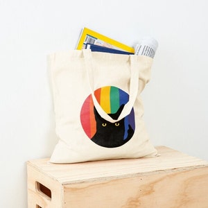 Op de afbeelding: Een witte canvas draagtas met een regenboogcirkel en een zwart katten silhouet erin. De kat kijkt de kijker aan met een ondeugende uitdrukking.
