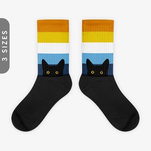 Puede incluir: Un par de calcetines negros con un patrón de rayas coloridas en la parte superior. Un gato negro con ojos amarillos asoma por detrás de las rayas.